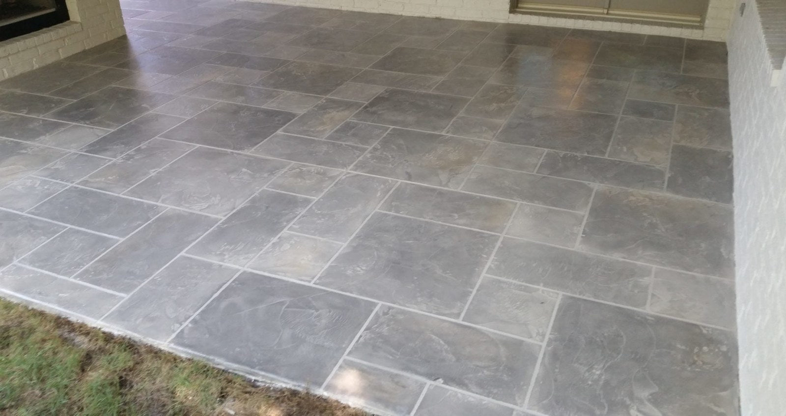 A covered residential patio finished with a grey concrete overlay troweled and grouted to look like large-format slate tile, with the edge of the original slab visible at the lawn